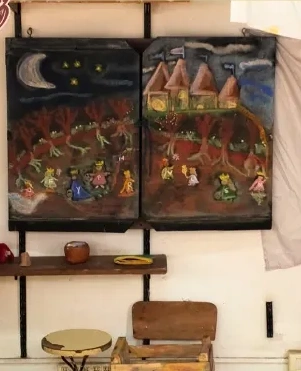 Pizarrón del aula con dibujo nocturno de tiza, estante con vasijas y sillita de madera